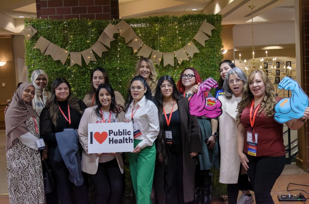 12 people pose for a photo in front of a green grass background. They are smiling and holding public health props like a sign that reads, I love public health.